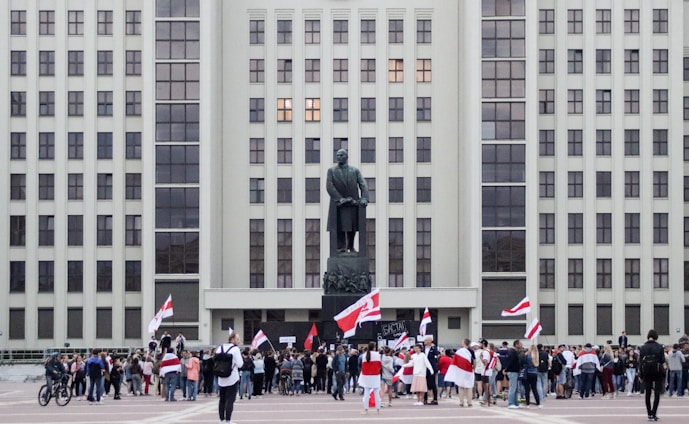 A large group of people gathers in front of a tall, rectangular government building with numerous windows. A statue stands prominently in front of the building, and many people in the crowd are holding red and white flags.