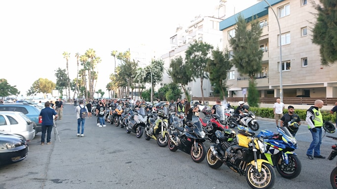 A group of motorcyclists gather on a city street, with a lineup of motorcycles parked along the road. The scene takes place in an urban area, with residential buildings and palm trees in the background. Several people stand near the motorcycles, engaging in conversation and preparing for a ride.