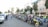 A group of motorcyclists gather on a city street, with a lineup of motorcycles parked along the road. The scene takes place in an urban area, with residential buildings and palm trees in the background. Several people stand near the motorcycles, engaging in conversation and preparing for a ride.