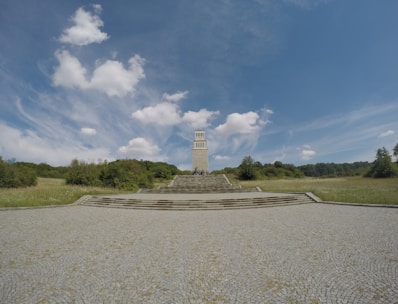 A tall stone tower stands at the center of a spacious landscape, surrounded by greenery and trees. The sky above is vast and blue, dotted with a few fluffy clouds, creating a serene and open atmosphere. The tower is accessed via several stone steps that lead up from a large cobblestone courtyard.