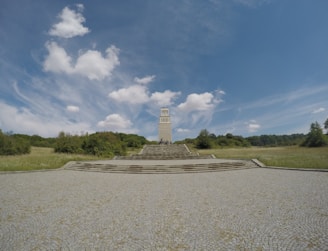 A tall stone tower stands at the center of a spacious landscape, surrounded by greenery and trees. The sky above is vast and blue, dotted with a few fluffy clouds, creating a serene and open atmosphere. The tower is accessed via several stone steps that lead up from a large cobblestone courtyard.