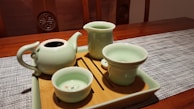 A wooden table set with a textured gray placemat holds a wooden tray featuring a set of light green ceramic teaware, including a teapot, a tall jug, and two cups. The teapot and cups have a glossy finish, and one cup has a small floral pattern inside. The scene is warmly lit, highlighting the smooth surfaces of the ceramics.