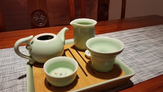 A wooden table set with a textured gray placemat holds a wooden tray featuring a set of light green ceramic teaware, including a teapot, a tall jug, and two cups. The teapot and cups have a glossy finish, and one cup has a small floral pattern inside. The scene is warmly lit, highlighting the smooth surfaces of the ceramics.