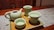 A wooden table set with a textured gray placemat holds a wooden tray featuring a set of light green ceramic teaware, including a teapot, a tall jug, and two cups. The teapot and cups have a glossy finish, and one cup has a small floral pattern inside. The scene is warmly lit, highlighting the smooth surfaces of the ceramics.
