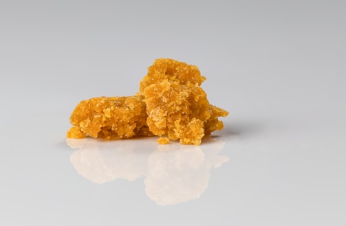 A pile of orange, granular substance rests on a smooth, reflective surface. The texture appears crumbly and rough, with irregular shapes throughout its form. The background is a clean, neutral tone, accentuating the vibrant color of the substance.