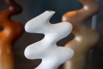The image features a set of abstract sculptures with smooth, undulating shapes. The forms are elegantly arranged, with a prominent white sculpture in the foreground and others in shades of brown and bronze in the background. The soft curves create a dynamic yet harmonious composition.