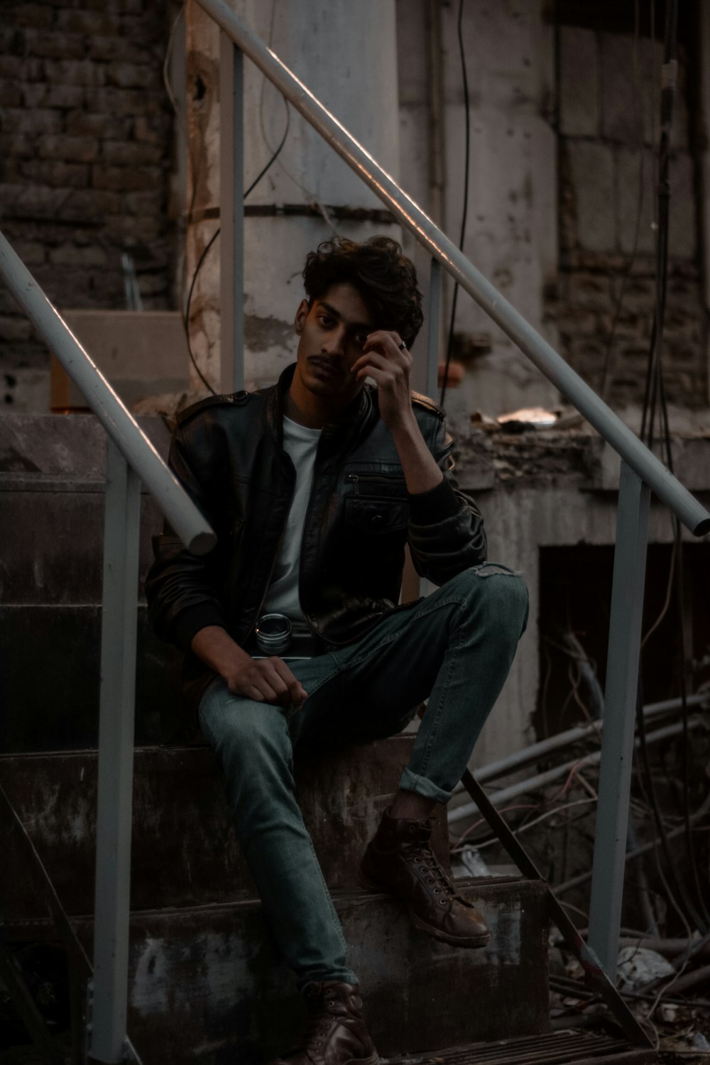 man in black leather jacket and blue denim jeans sitting on gray metal stair