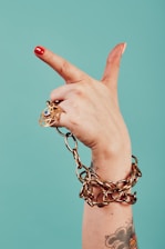 A hand with red nail polish is holding a large gold chain. The hand is adorned with a gold ring featuring a blue gem, and there is a tattoo visible on the wrist. The background is a solid teal color.