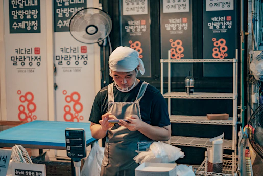 A man wearing an apron and headscarf is focused on his phone, standing in what seems to be a small shop or stall. Behind him, there are shelves and signs with Korean text. The area is cluttered with various shop items like a fan, scale, and bins.