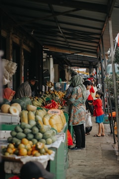 A bustling market scene with various tropical fruits displayed on the stall, including melons, bananas, and papayas. Several people are engaged in shopping and interacting with vendors under a covered walkway. The atmosphere is lively and casual.