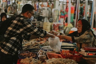 A man wearing a mask and a checkered shirt is purchasing items from a street market vendor, who is an older woman wearing a headscarf. The vendor hands over products wrapped in plastic bags, while the counter is filled with a variety of unpackaged food items.