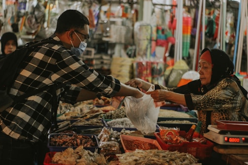 A man wearing a mask and a checkered shirt is purchasing items from a street market vendor, who is an older woman wearing a headscarf. The vendor hands over products wrapped in plastic bags, while the counter is filled with a variety of unpackaged food items.