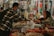 A man wearing a mask and a checkered shirt is purchasing items from a street market vendor, who is an older woman wearing a headscarf. The vendor hands over products wrapped in plastic bags, while the counter is filled with a variety of unpackaged food items.
