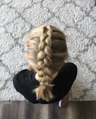 A person with blonde hair styled in a neat French braid, sitting on a patterned carpet. The individual is wearing a black top and leaning slightly forward, with the braid clearly visible from above.