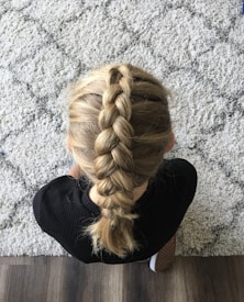 A person with blonde hair styled in a neat French braid, sitting on a patterned carpet. The individual is wearing a black top and leaning slightly forward, with the braid clearly visible from above.