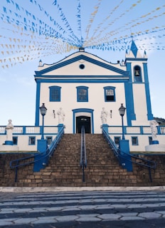 A white and blue church stands prominently with a set of stone stairs leading up to it. Colorful triangular flags are strung across the sky, adding a festive atmosphere. Sculptures are positioned along the stairway, and two vintage-style lamps are placed near the entrance.