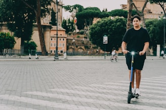 A person is riding an electric scooter on a cobblestone street. The background features an urban setting with trees, buildings, and a few other people, one on a bicycle. The scene appears to be in a historical area with older architectural elements.