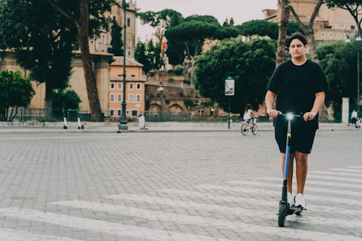 A person is riding an electric scooter on a cobblestone street. The background features an urban setting with trees, buildings, and a few other people, one on a bicycle. The scene appears to be in a historical area with older architectural elements.