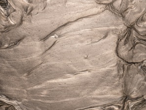 Swirling and smoothed texture of a muddy or clay-like substance, featuring a range of brown shades with some lighter streaks. The surface appears wet, with wavy patterns that suggest movement or layering of the material.