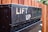 A close-up of a black metallic surface with water droplets and the text 'LIFT UP' prominently displayed. Behind it is a wooden fence, slightly out of focus.