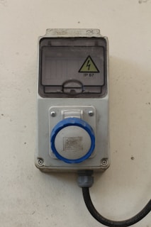 A wall-mounted industrial electrical socket featuring a circular blue plug and a protective cover marked with a high voltage warning sign labeled IP 67. The housing appears sturdy, made of a gray metallic or plastic material with a cable extending downward.