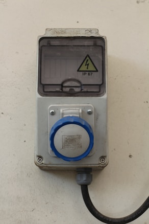 A wall-mounted industrial electrical socket featuring a circular blue plug and a protective cover marked with a high voltage warning sign labeled IP 67. The housing appears sturdy, made of a gray metallic or plastic material with a cable extending downward.