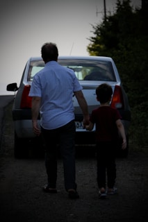 A man and a child are walking hand in hand toward a parked car on a rural road. The setting appears to be evening or early morning with subdued lighting. The man is wearing a striped shirt and dark pants, while the child is dressed in a dark t-shirt and dark pants. They are moving away from the camera, adding a sense of journey or departure.