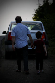 A cheerful dad holding a toddler’s hand by a roadside with a thumb out, ready to hitchhike.