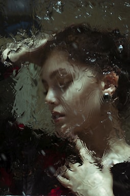 A portrait of a person seen through a textured glass or water surface, creating a distorted and artistic effect. The person has their eyes closed and a serene expression. They are touching their face and appear to be in a moment of contemplation or relaxation.