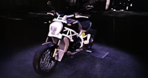 A sleek and modern motorcycle is parked on a dimly lit pavement. The motorcycle features a distinctive white frame with black accents, and it includes elements such as a visible exhaust and shiny spokes on the wheels. Part of a dark, industrial background is visible, which enhances the focus on the motorcycle.
