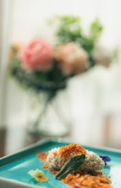 A close-up of a gourmet dish presented on a blue plate, featuring a textured topping resembling breadcrumbs, garnished with a green leaf and small edible flowers. In the blurred background, there is a vase with pastel-colored flowers.