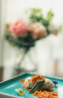 A close-up of a gourmet dish presented on a blue plate, featuring a textured topping resembling breadcrumbs, garnished with a green leaf and small edible flowers. In the blurred background, there is a vase with pastel-colored flowers.