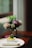 A white rectangular dish filled with sliced ginger and garnished with herbs and purple flowers sits on a white ceramic plate. Beside the dish, there is a rolled napkin. In the background, out of focus, is a vase holding a bouquet of pink and white flowers. The setting appears to be on a wooden table near a large window, allowing natural light to illuminate the scene.