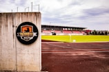 A soccer stadium with red and white bleachers is visible in the background, surrounded by a well-maintained green field. In the foreground, there is a concrete wall with a circular emblem that features a graphic design and the text 'FC'. The tracks around the field appear freshly paved, and the sky is overcast with gray clouds.
