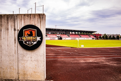 A soccer stadium with red and white bleachers is visible in the background, surrounded by a well-maintained green field. In the foreground, there is a concrete wall with a circular emblem that features a graphic design and the text 'FC'. The tracks around the field appear freshly paved, and the sky is overcast with gray clouds.