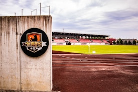 A soccer stadium with red and white bleachers is visible in the background, surrounded by a well-maintained green field. In the foreground, there is a concrete wall with a circular emblem that features a graphic design and the text 'FC'. The tracks around the field appear freshly paved, and the sky is overcast with gray clouds.
