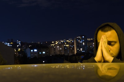 A person wearing a hood is clasping their hands together in a prayer-like gesture against the backdrop of a cityscape at night. The city lights are glowing in the distance, and the surface in front of the person reflects some of the light, adding to the serene atmosphere.