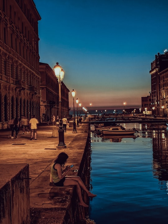 people walking on sidewalk near river during night time in Trieste Italy