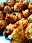 A close-up view of golden-brown fritters, possibly pakoras or bhajis, with a crispy texture and specks of herbs throughout. They are arranged on a metallic surface, indicating they may be freshly cooked.