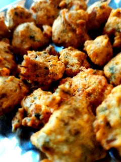 A close-up view of golden-brown fritters, possibly pakoras or bhajis, with a crispy texture and specks of herbs throughout. They are arranged on a metallic surface, indicating they may be freshly cooked.