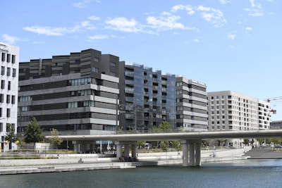 Several modern office buildings with glass and concrete facades line a waterfront. A bridge extends across the water, and there are people walking beneath and on the bridge. The scene is set against a blue sky with light clouds.