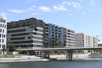 Several modern office buildings with glass and concrete facades line a waterfront. A bridge extends across the water, and there are people walking beneath and on the bridge. The scene is set against a blue sky with light clouds.