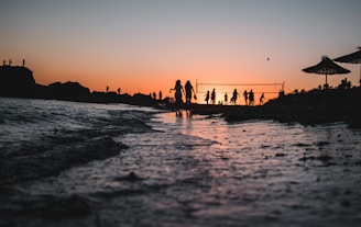 Silhouetted figures of people are playing beach volleyball near the shore during a beautiful sunset. The colors of the setting sun create a stunning gradient across the sky, reflecting off the water. There are a few parasols visible, and the shoreline is dotted with gentle waves.