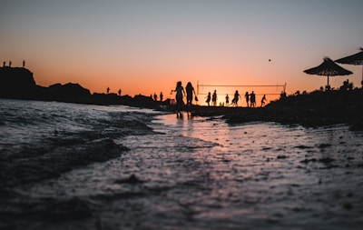 Silhouetted figures of people are playing beach volleyball near the shore during a beautiful sunset. The colors of the setting sun create a stunning gradient across the sky, reflecting off the water. There are a few parasols visible, and the shoreline is dotted with gentle waves.