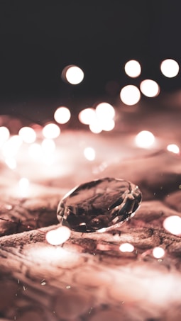 A glistening diamond sits atop a textured surface, surrounded by an array of defocused warm lights, creating a bokeh effect. The diamond is the focal point, reflecting subtle sparkles.