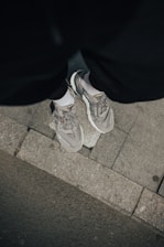 A person wearing gray running shoes stands on a sidewalk with a rough textured surface. The shoes are paired with black pants and white socks, and the ground is composed of both concrete and asphalt.