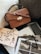 A brown leather handbag with gold hardware rests on a textured gray cushion. Below the bag, a collection of fashion magazines and a pair of black glasses are arranged next to a laptop. The scene is set against a white, fluffy surface, creating a chic and stylish composition.