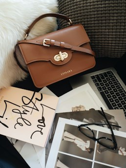 A brown leather handbag with gold hardware rests on a textured gray cushion. Below the bag, a collection of fashion magazines and a pair of black glasses are arranged next to a laptop. The scene is set against a white, fluffy surface, creating a chic and stylish composition.
