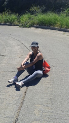 A person wearing athletic clothing and a cap is sitting on a paved surface with greenery in the background. They appear to be outdoors, sitting in a relaxed posture next to a red bag.