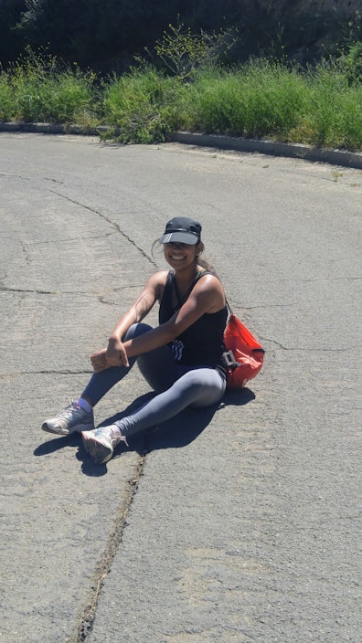 A person wearing athletic clothing and a cap is sitting on a paved surface with greenery in the background. They appear to be outdoors, sitting in a relaxed posture next to a red bag.